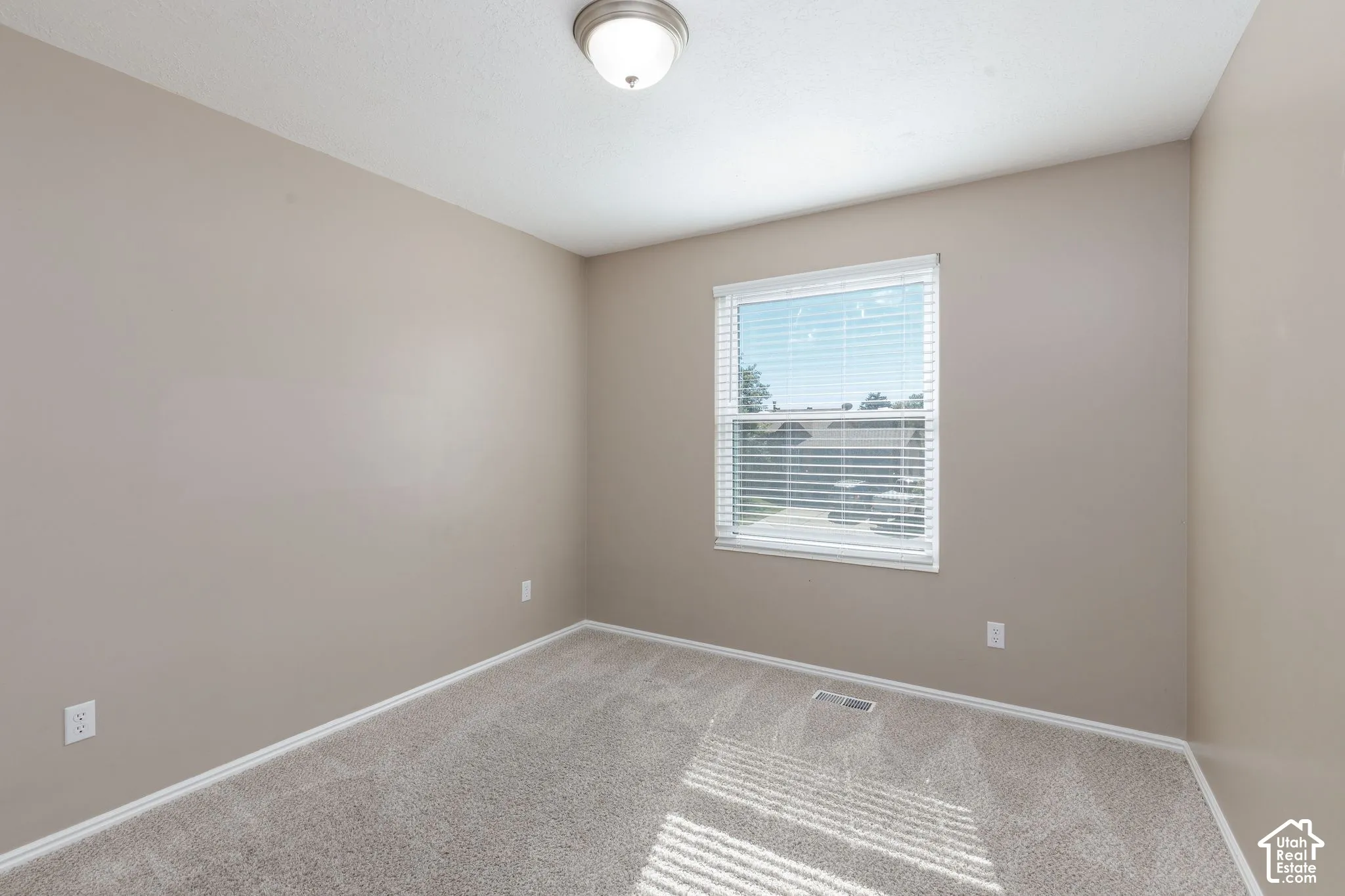 Neutral-toned walls and plush carpeting provide a versatile space, complemented by a single window with blinds and a ceiling-mounted light fixture
