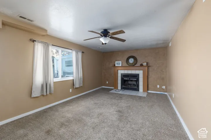 Carpeted room featuring a ceiling fan, fireplace with tile surround, wood mantel, and window with white trim