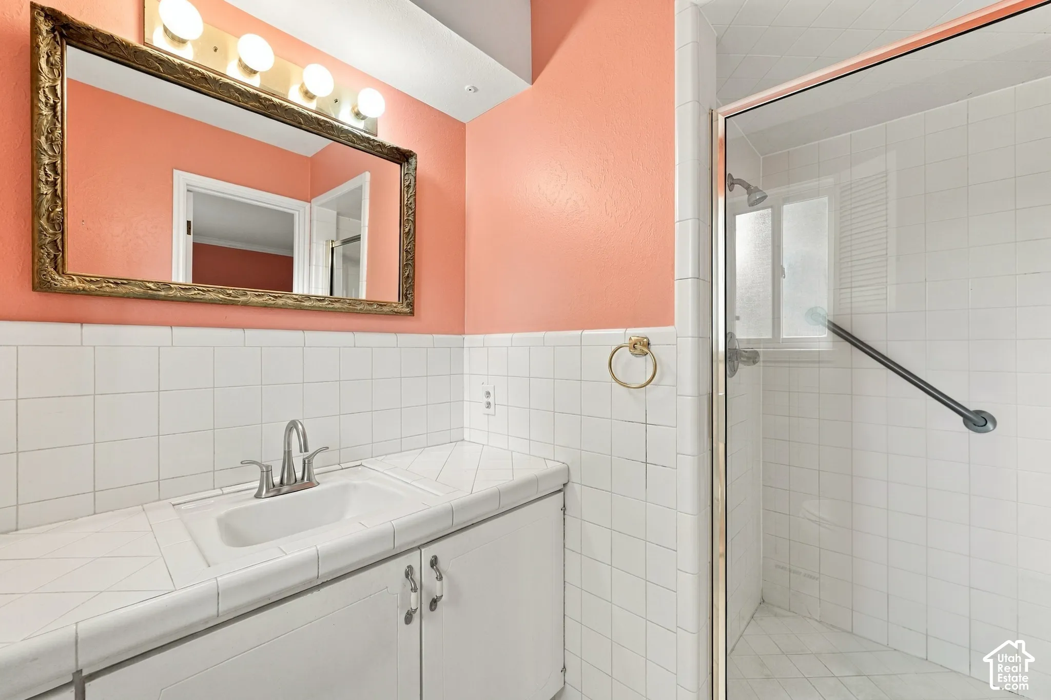 Ensuite Bathroom vanity featuring a tile countertop with an integrated basin, white cabinetry, a framed rectangular mirror, four-bulb vanity lighting, and a glass-enclosed shower with tiled surround