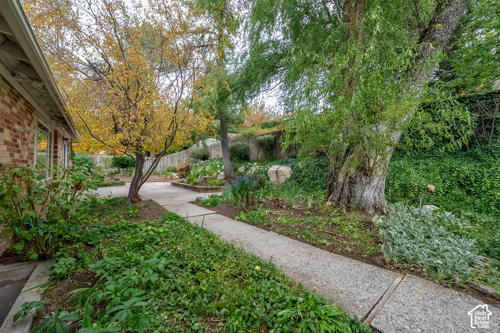Exterior rear yard featuring a concrete patio, retaining wall, mature trees, tiered garden beds, and brick home siding
