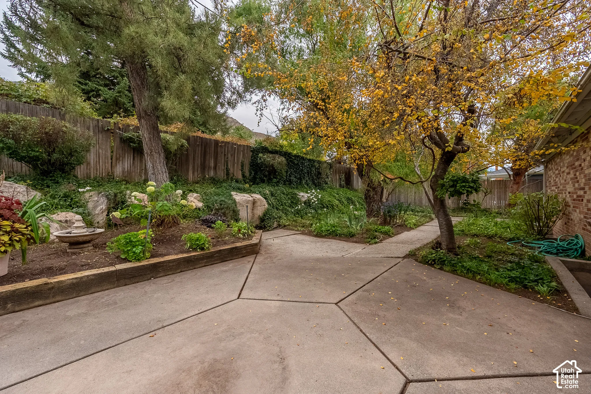 Rear grounds featuring concrete patio surfacing, tiered garden beds with wood-timber retaining walls, mature trees, and rock-faced landscaping