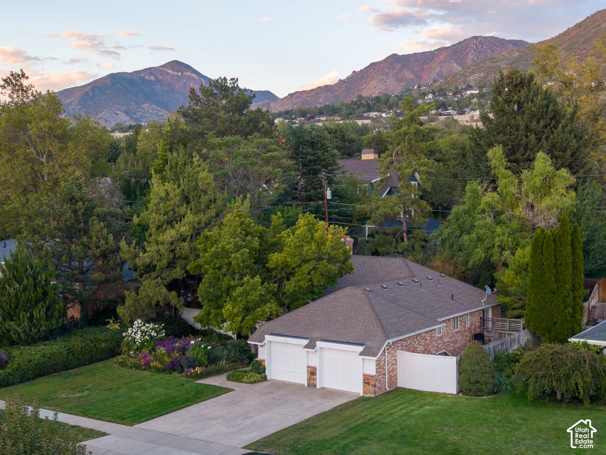Mountain backdrop, mature trees, landscaped garden beds, manicured lawn, attached two-car garage