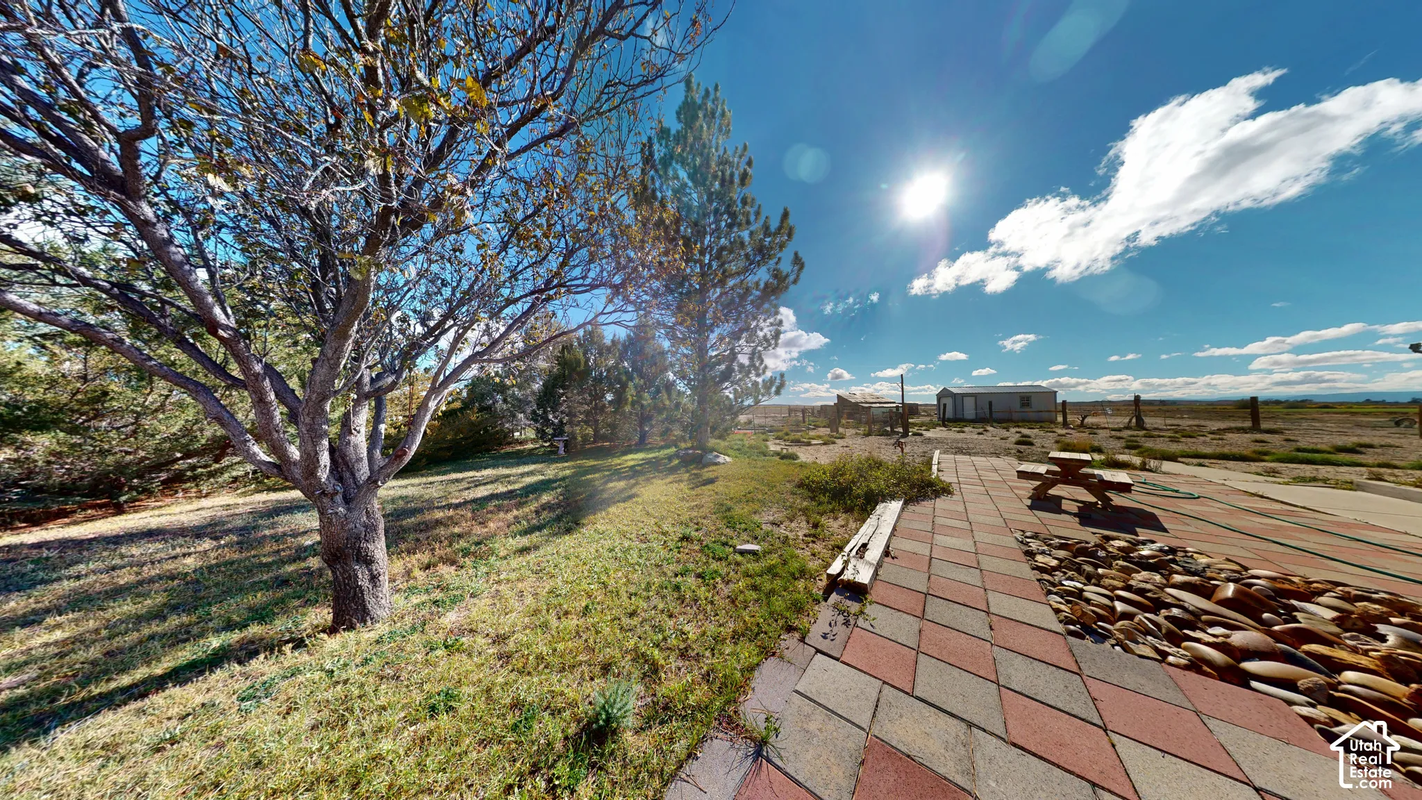 Outdoor living area featuring a paved multi-tonal patio, decorative stone bed, mature trees, and open terrain