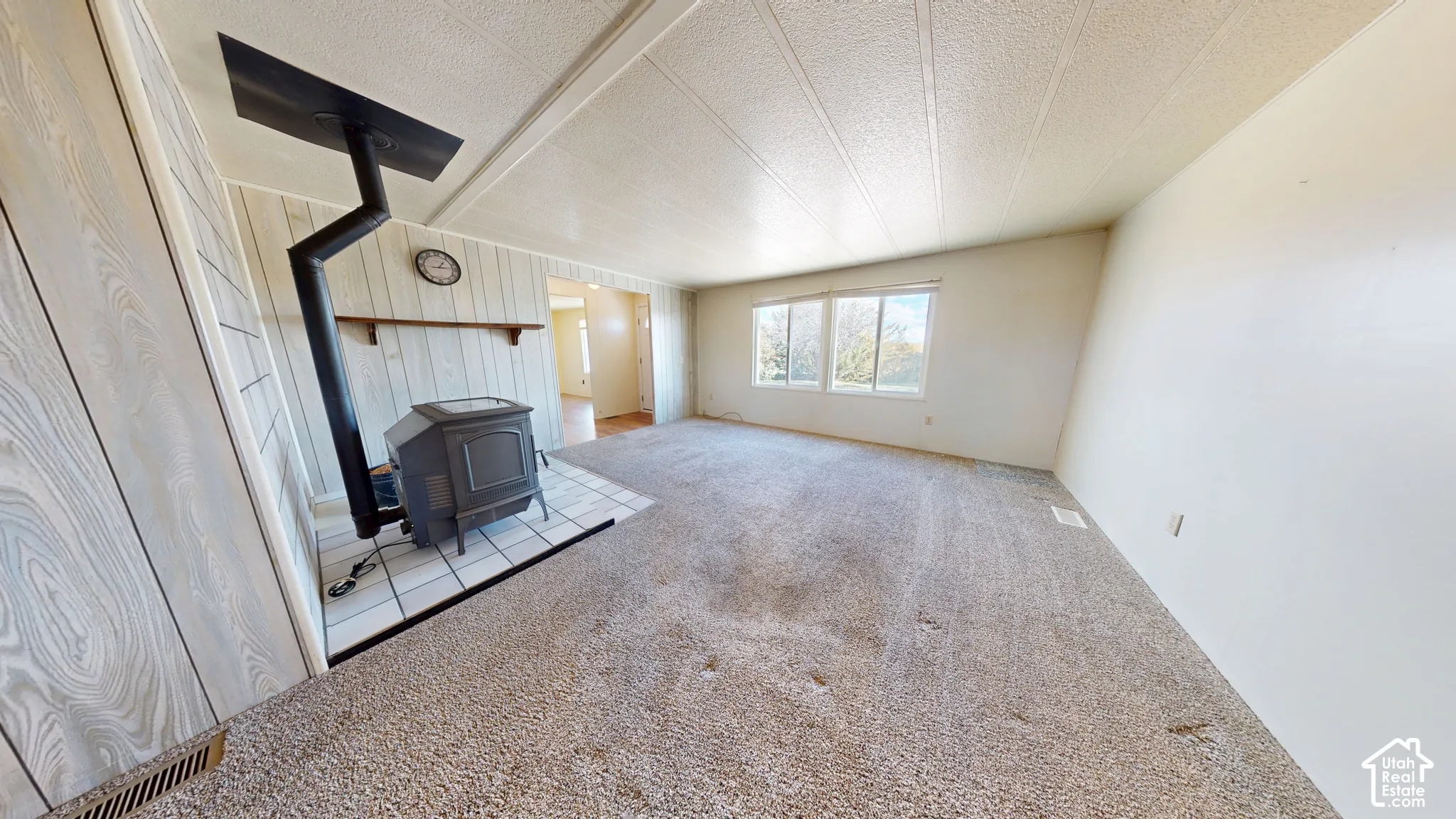 Living area featuring wood-paneled walls, a standalone metal wood-burning stove on a tiled hearth, dual windows, and wall-to-wall carpeting