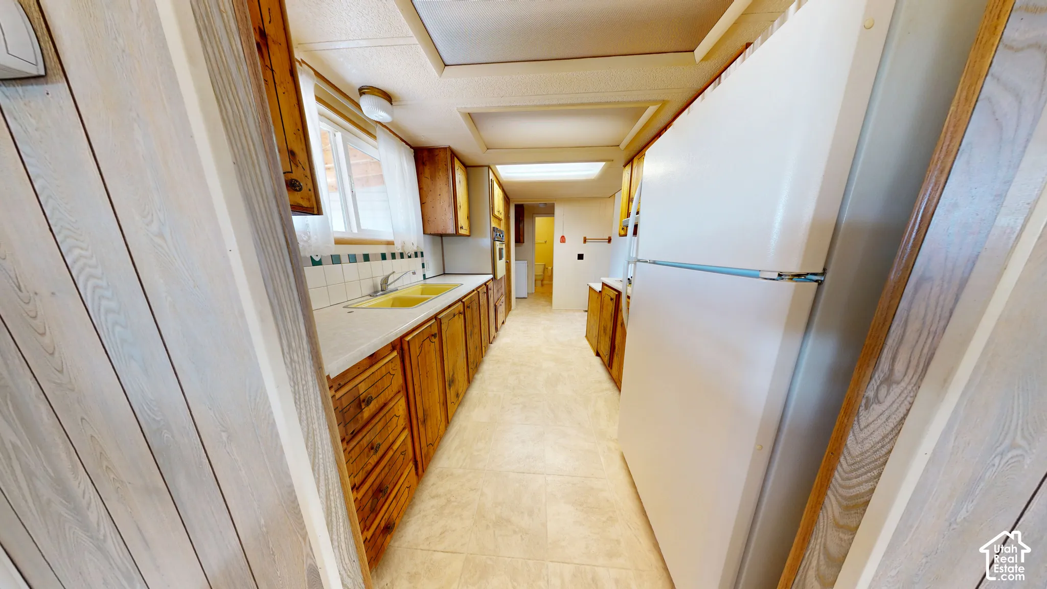 Galley kitchen featuring natural wood cabinetry, white countertops, square tile backsplash, integrated lighting, and neutral-tone flooring