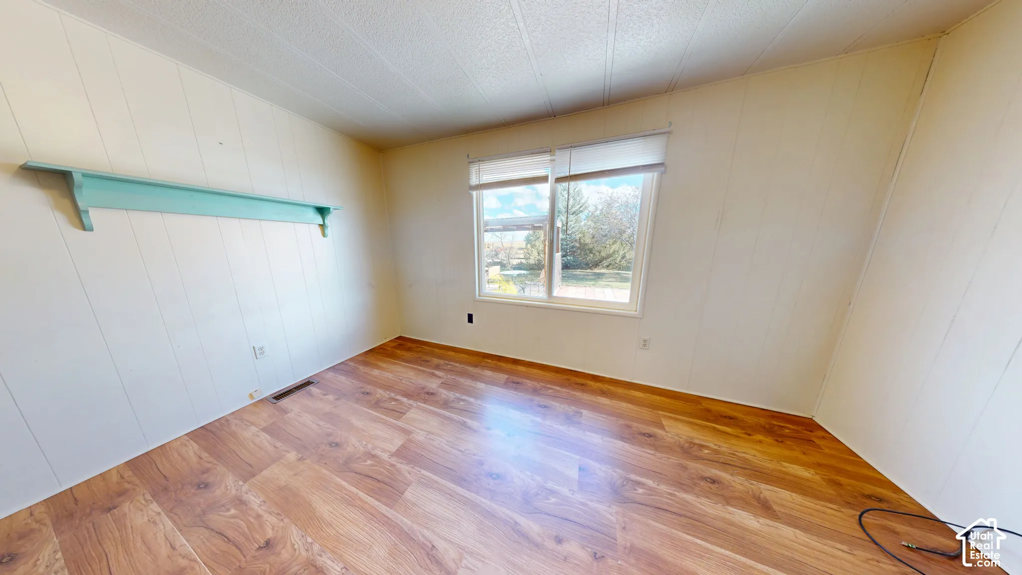Interior room featuring wood-finish flooring, panelled walls, a large window with fitted roller shade, and a built-in wall-mounted shelf