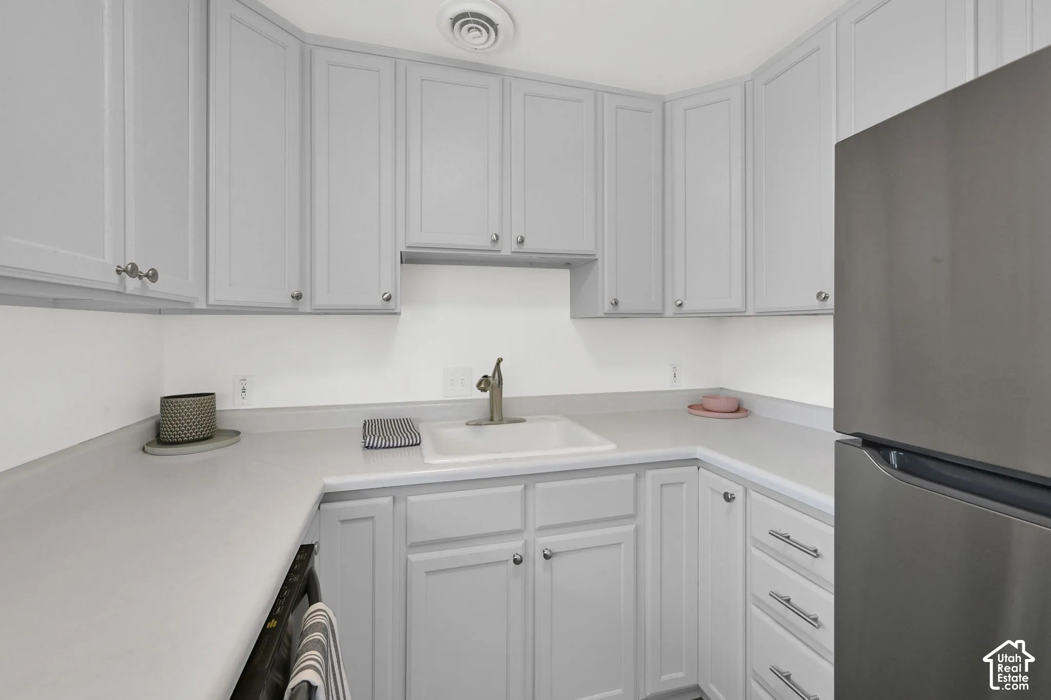 U-shaped kitchen featuring light gray cabinetry with silver hardware, light gray countertops, a single basin sink with a bronze faucet, and a stainless steel refrigerator