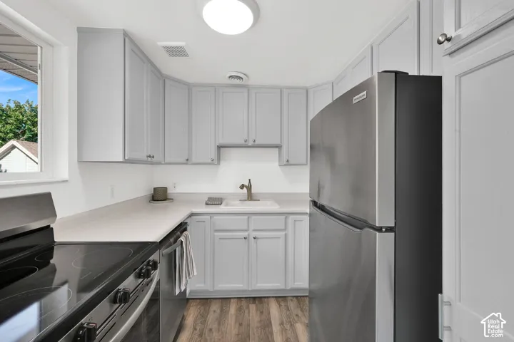 Light gray cabinetry, stainless steel appliances, light-toned countertops, wood-finish flooring, and a white sink with a bronze-toned faucet