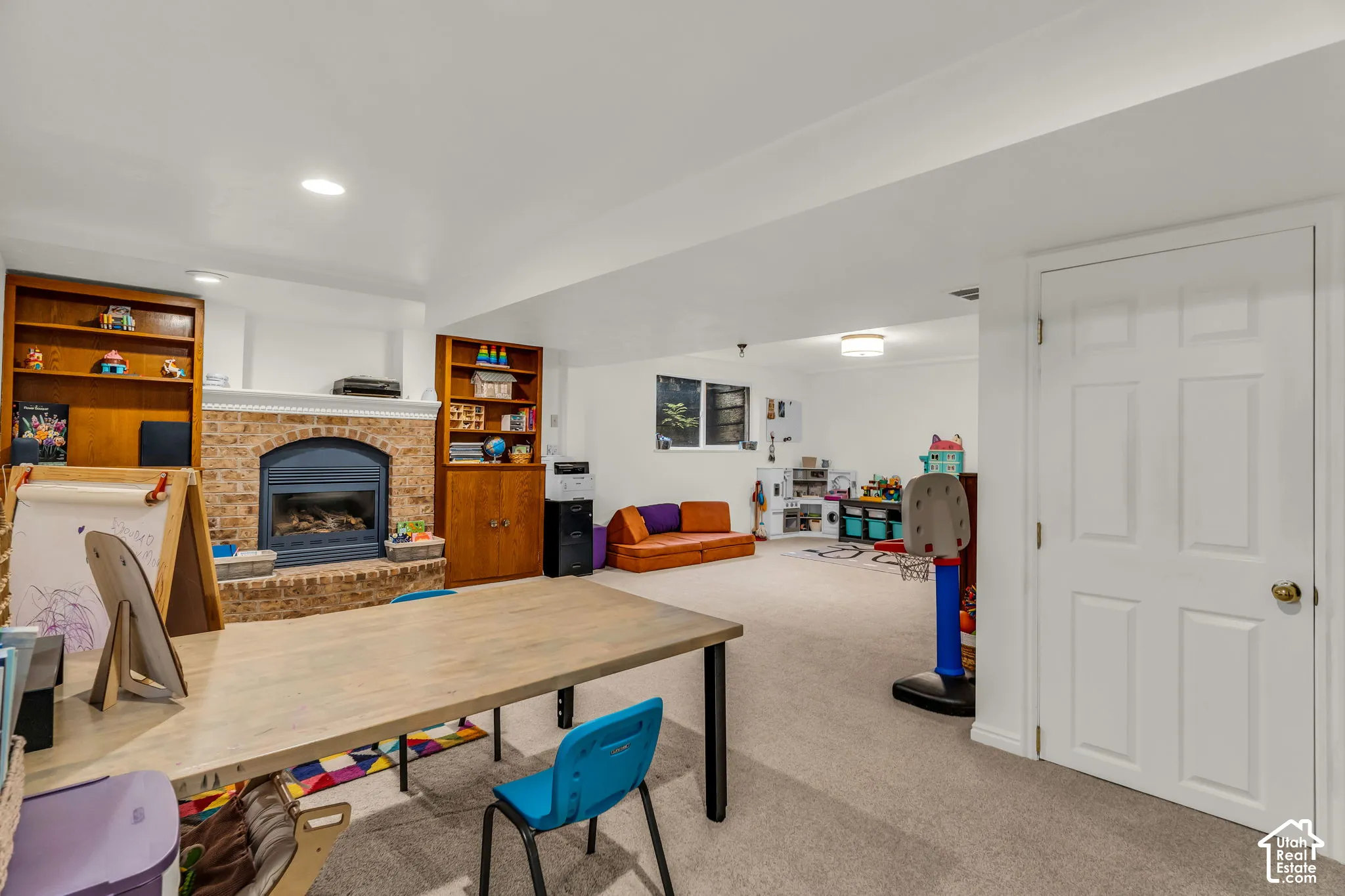 Spacious room featuring a brick-surround fireplace with a dark metal insert, built-in wood shelving, recessed lighting, and light-colored carpeting