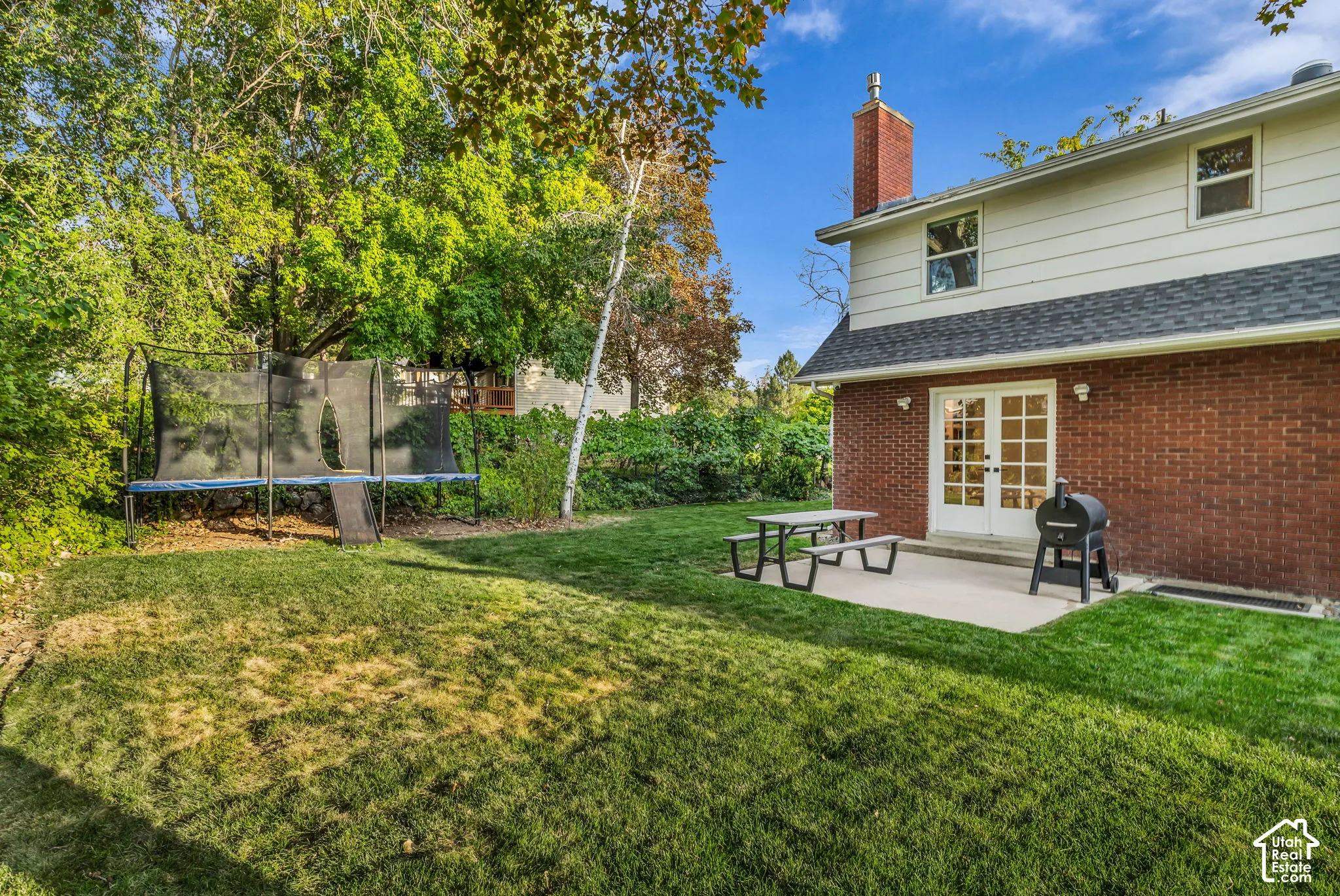 Expansive backyard featuring a manicured lawn, established perimeter trees, a concrete patio, and white French doors
