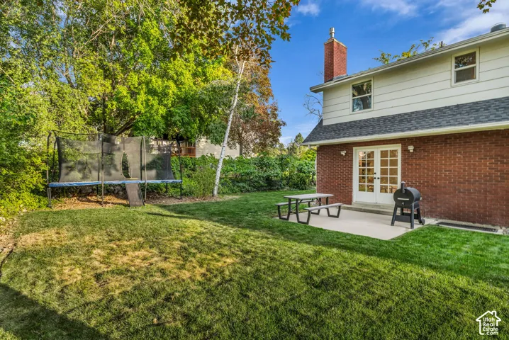 Expansive backyard featuring a manicured lawn, established perimeter trees, a concrete patio, and white French doors