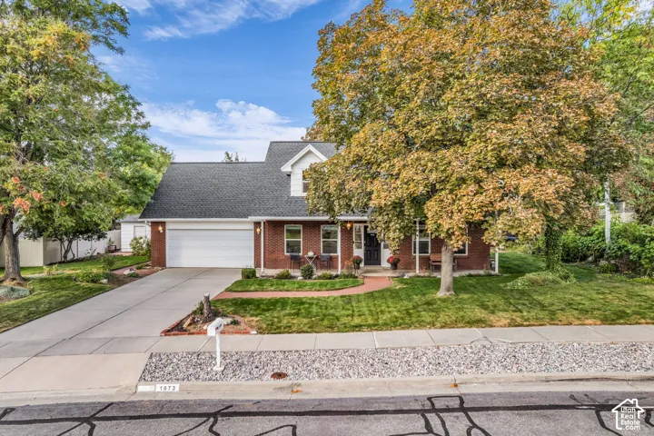 Brick exterior with a white garage door, gabled roof, mature trees, concrete driveway, and a manicured lawn