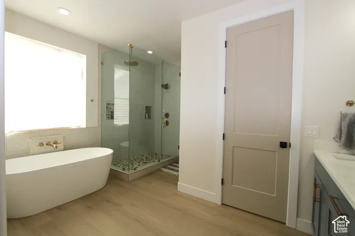 Freestanding soaking tub, glass-enclosed shower with pebble flooring, light wood-finish flooring, and a vanity with a white countertop