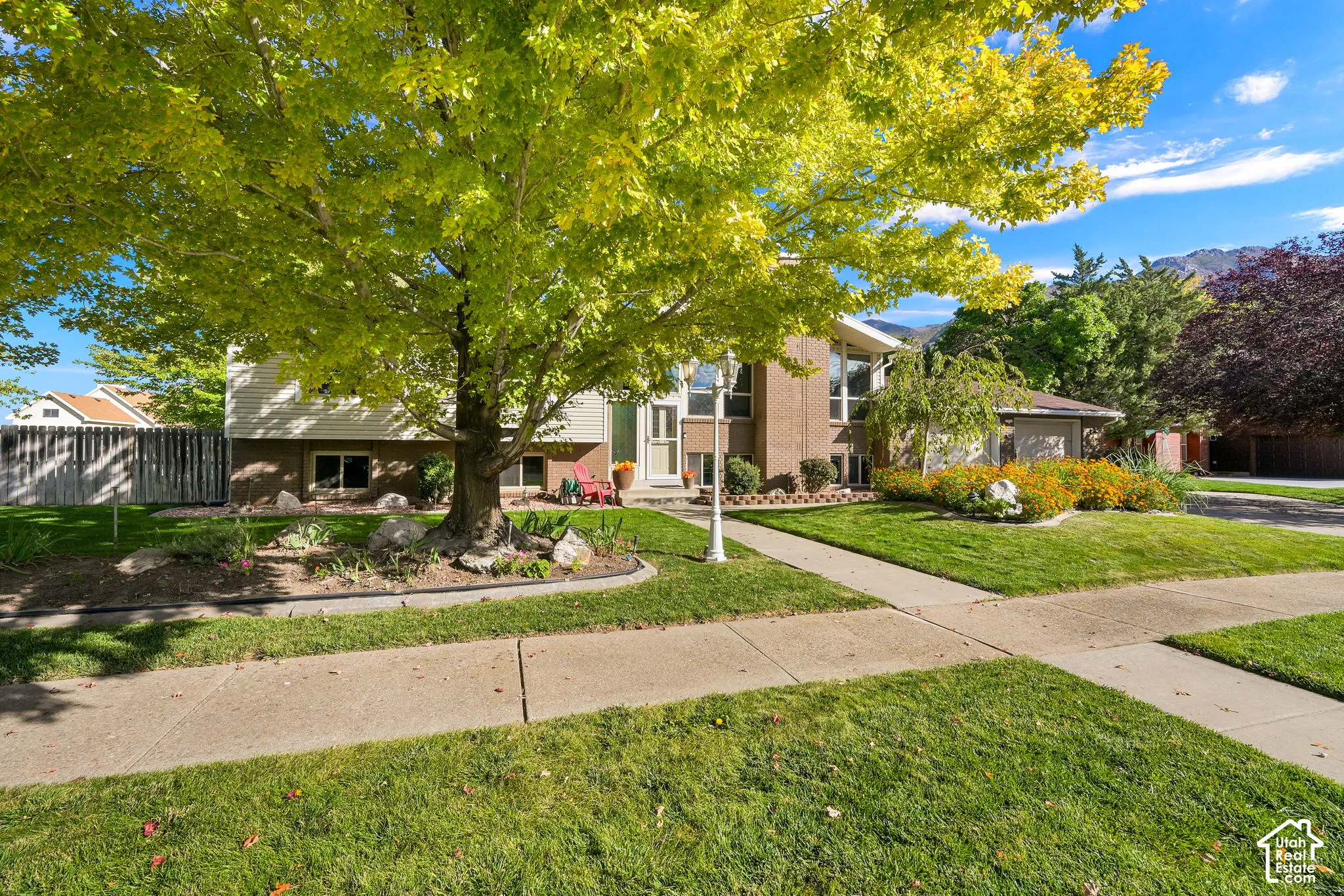 Brick exterior with vertical siding, mature trees, concrete sidewalks, and landscaped garden beds