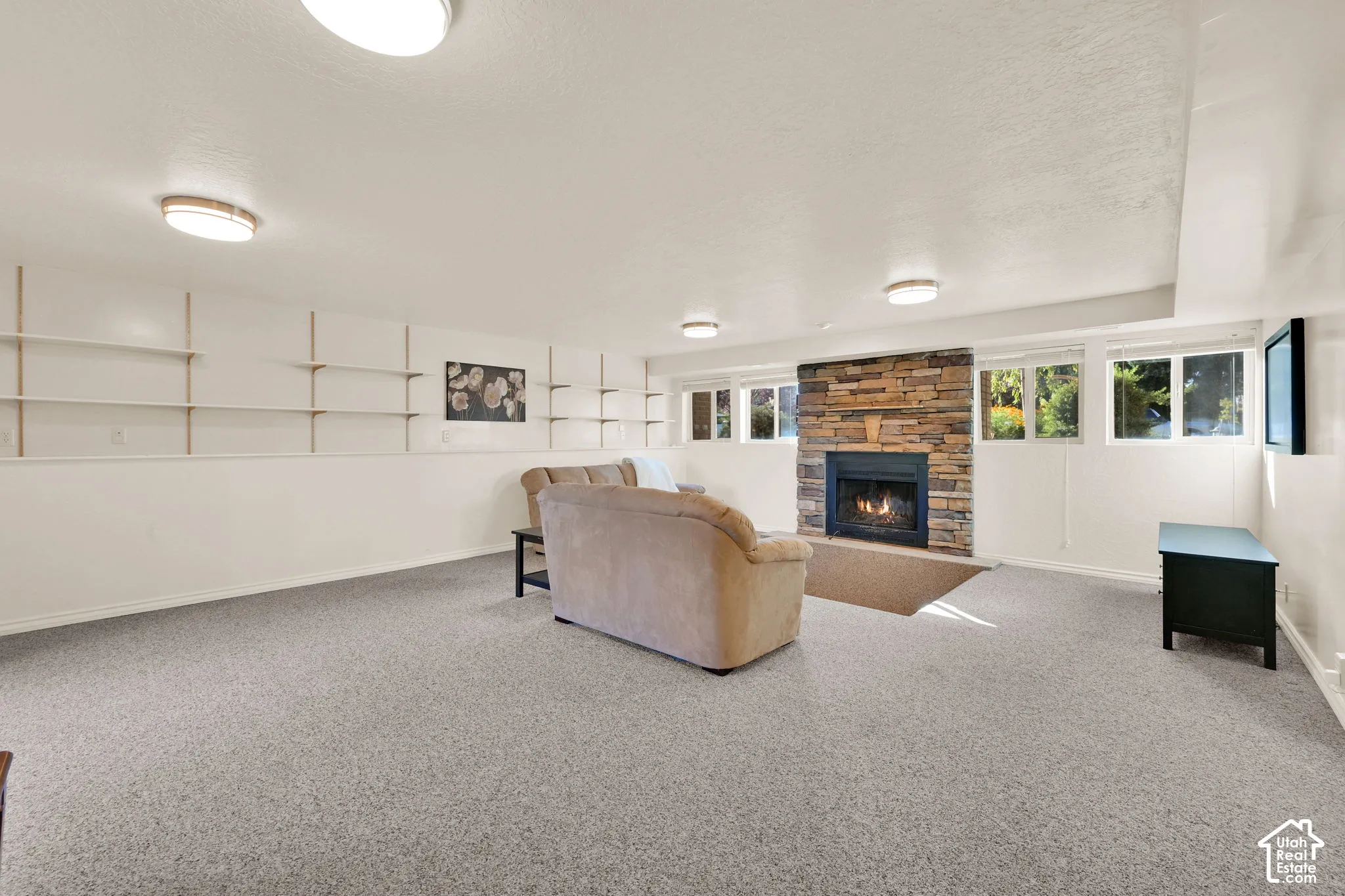 Spacious basement living room featuring light-toned carpeting, a stone-faced fireplace with built-in shelving, white walls, and multiple windows