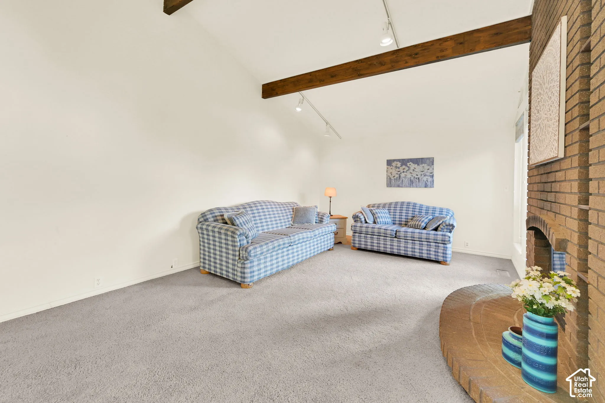 Vaulted ceiling with exposed wood beams, brick fireplace with a curved hearth, track lighting, and light-toned carpeting