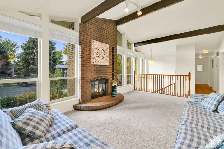 Vaulted ceiling with exposed wood beams, brick fireplace with hearth, wall of windows, and carpeted flooring