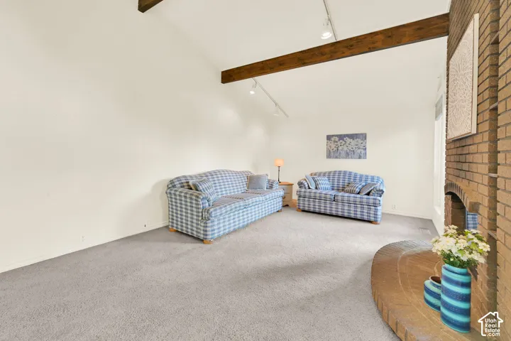 Vaulted ceiling with exposed wood beams, brick fireplace with a curved hearth, track lighting, and light-toned carpeting