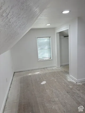 Sloped ceiling with textured finish, recessed lighting, window with blinds, baseboard trim, and wood plank flooring