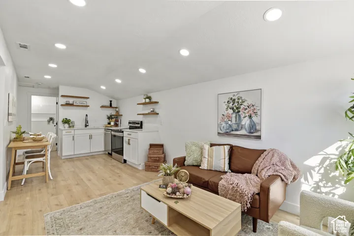 Open-concept living and kitchen space featuring recessed ceiling lighting, wood-finish flooring, white cabinetry with stainless steel appliances, floating wooden shelves, and a leather seating area with natural light from expansive windows