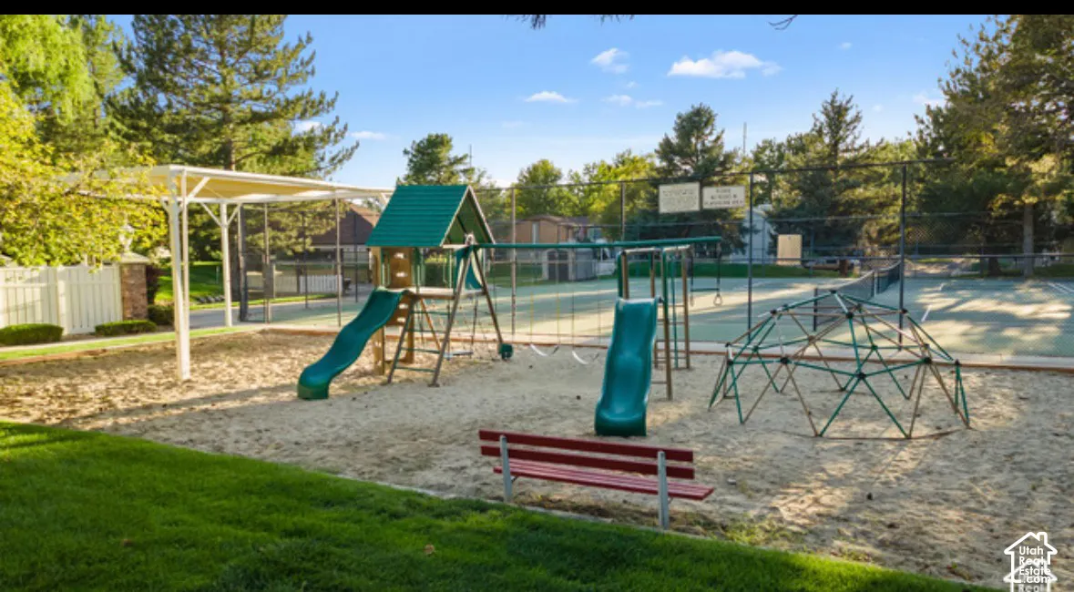 Recreational area featuring a sand-surface playground with a climbing structure, slides, a tennis court, and a shaded pavilion