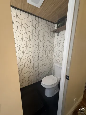 Geometric patterned wallpaper, wood-finish ceiling paneling, white dual-flush toilet, and wall-mounted floating shelf