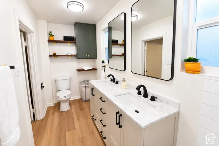 Dual-sink vanity with a solid-surface countertop and matte black fixtures, custom cabinetry, wood-finish flooring, floating wood shelving, and rectangular wall-mounted mirrors