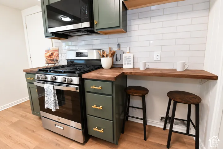 Kitchen featuring sage green cabinetry with brass hardware, wood-finish countertops, stainless steel gas range, over-the-range microwave, and white subway tile backsplash