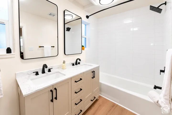 Dual-sink vanity with a stone-finish countertop, matte black fixtures, rectangular undermount sinks, white subway-tiled shower surround, and wood-finish flooring
