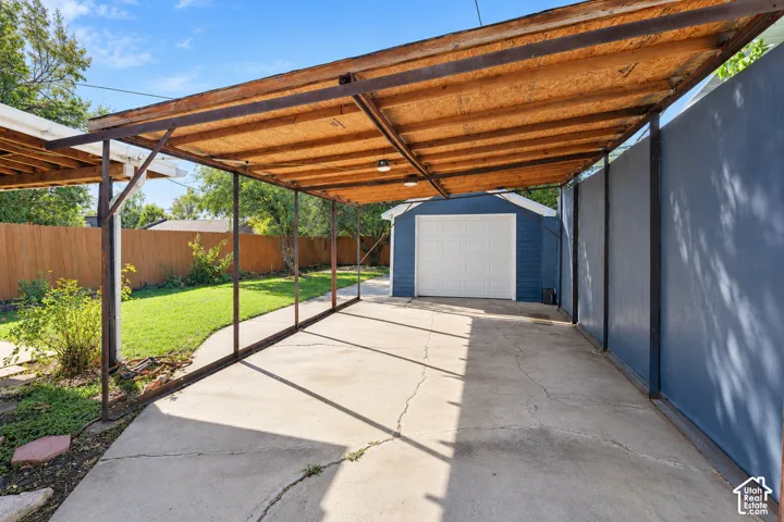 Covered carport featuring a structural steel frame with wood-sheathed roofing, concrete paving, and an attached detached garage with a white roll-up door