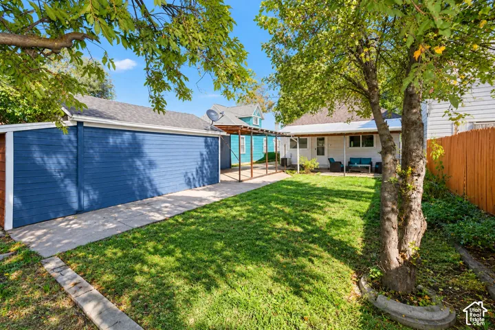 Rear yard featuring a manicured lawn, a detached garage with blue horizontal siding, a concrete patio, a covered pergola, and mature trees