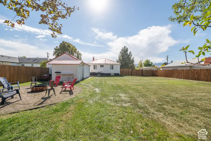 Expansive fenced backyard featuring a large grass area, a dedicated fire pit area, and a white auxiliary structure with a red metal roof