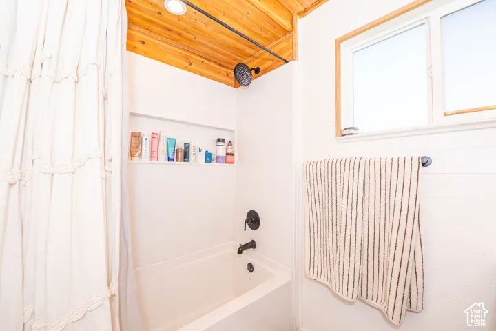White bathtub with matte black fixtures, an integrated shower niche, and a natural wood-finish ceiling