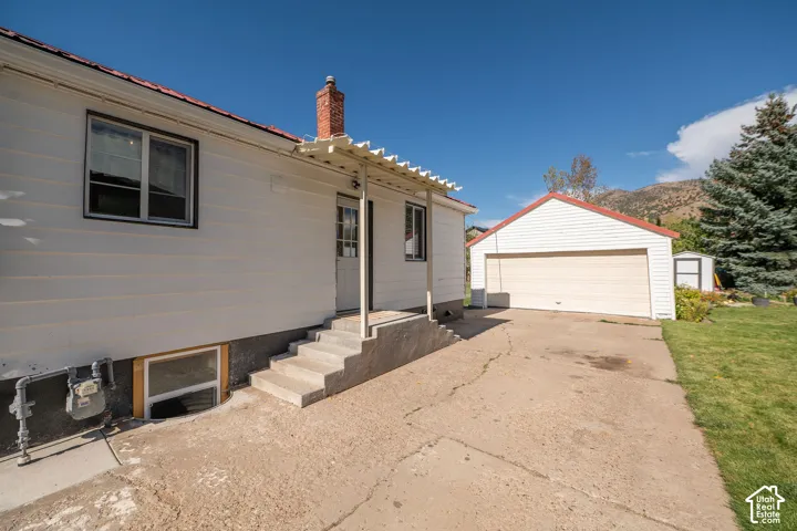 Detached two-car garage, concrete driveway, concrete exterior steps, brick chimney, and slatted patio cover