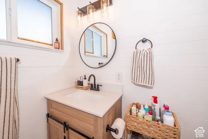 White shiplap wall paneling, vanity with a light-toned wood finish and barn door-style cabinet, integrated sink with a light countertop, round black-framed mirror, and a three-light vanity fixture with clear glass shades