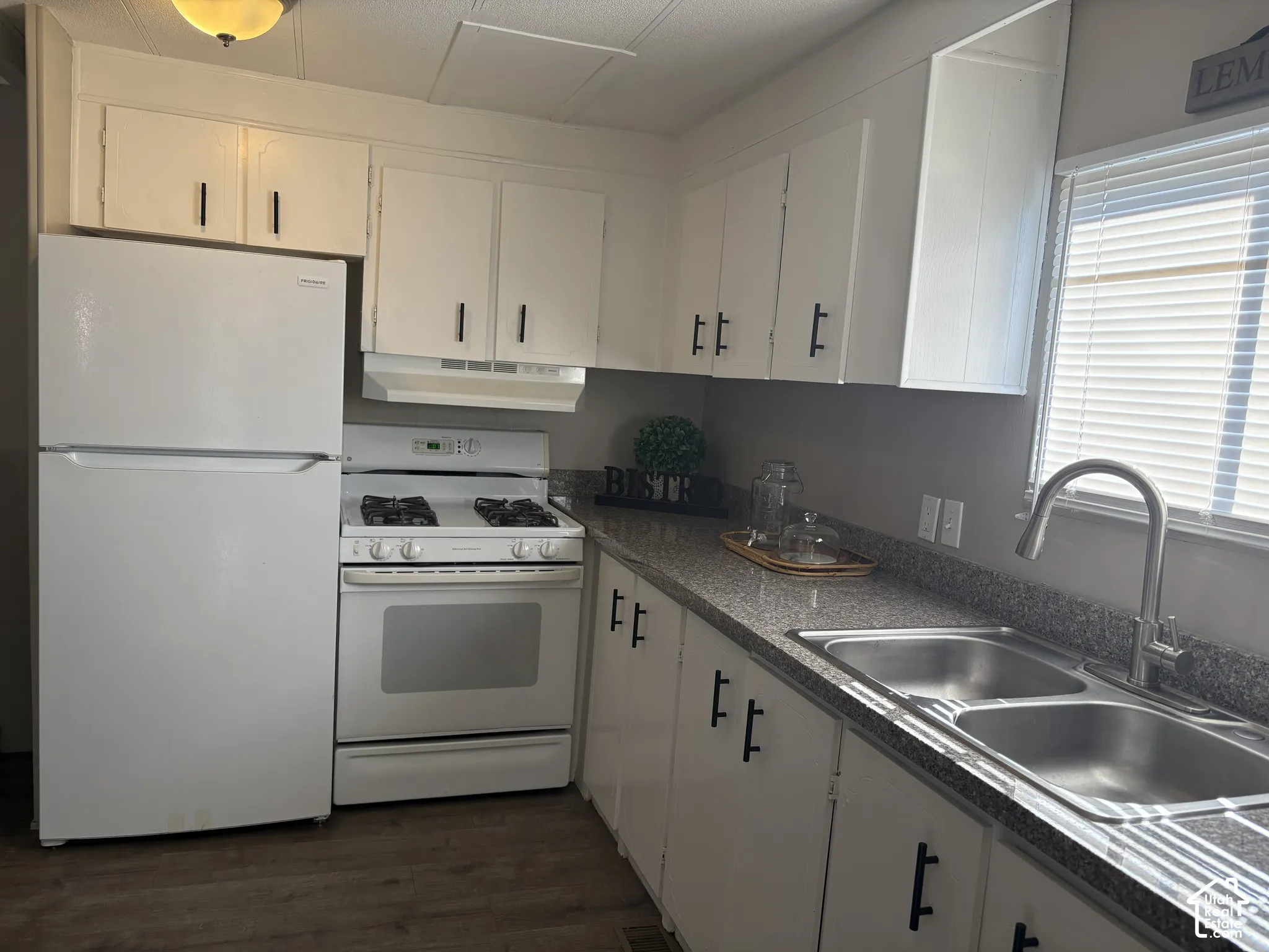 White cabinetry with matte black hardware, dual basin stainless steel sink, wood-finish flooring, and a white appliance package