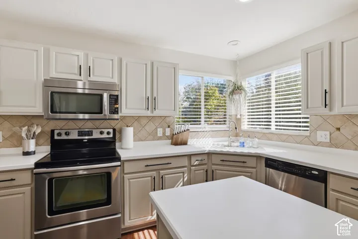 White cabinetry with black hardware, stainless steel appliances, white countertops, and a tiled backsplash, featuring a corner sink with windows above