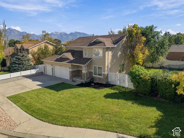 Stucco exterior with stone accents, two-car garage, manicured lawn, white picket fence, and mountain backdrop