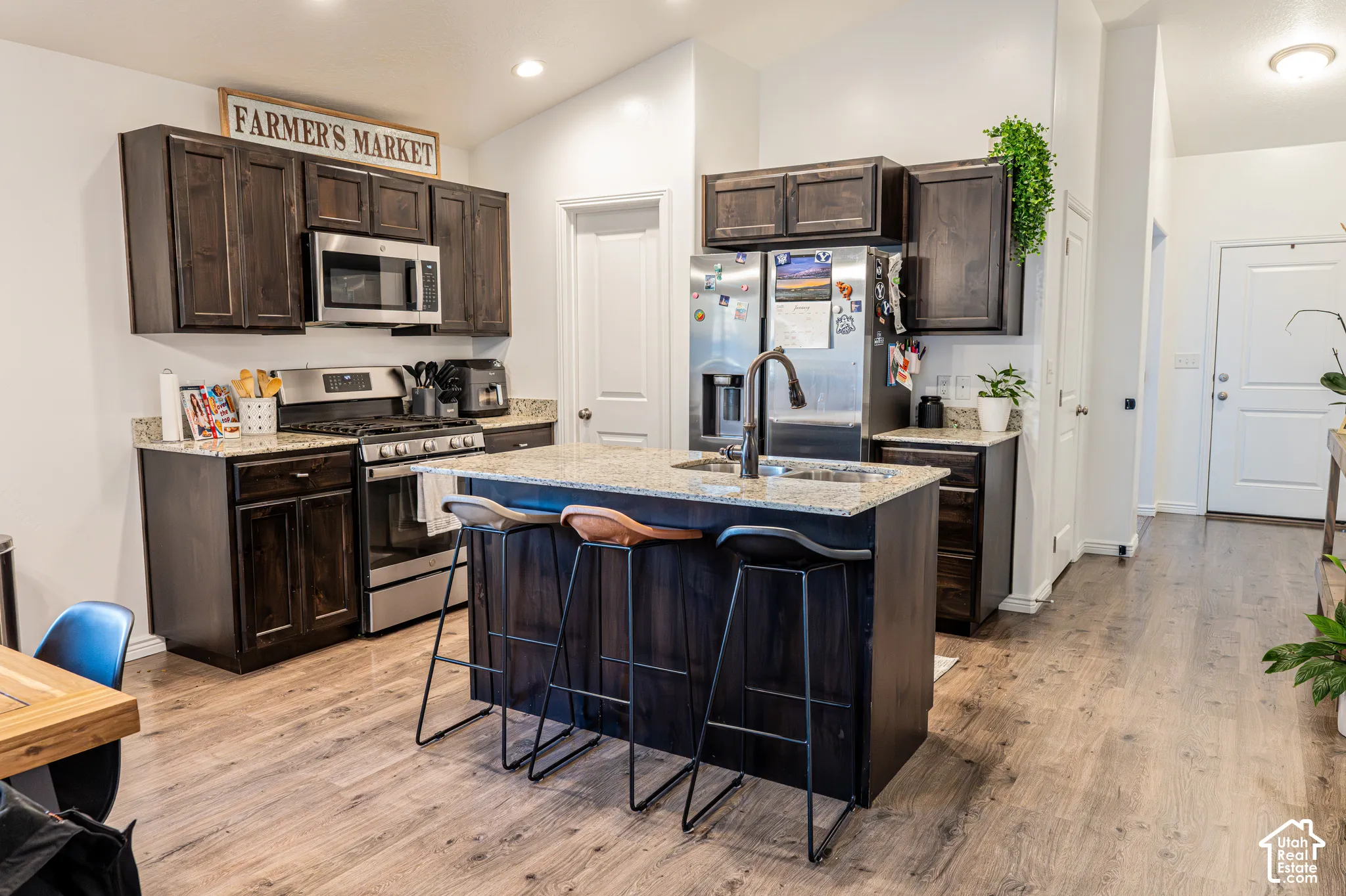 Kitchen featuring dark wood cabinetry, granite countertops, stainless steel appliances, a center island with a built-in sink, and wood-finish flooring