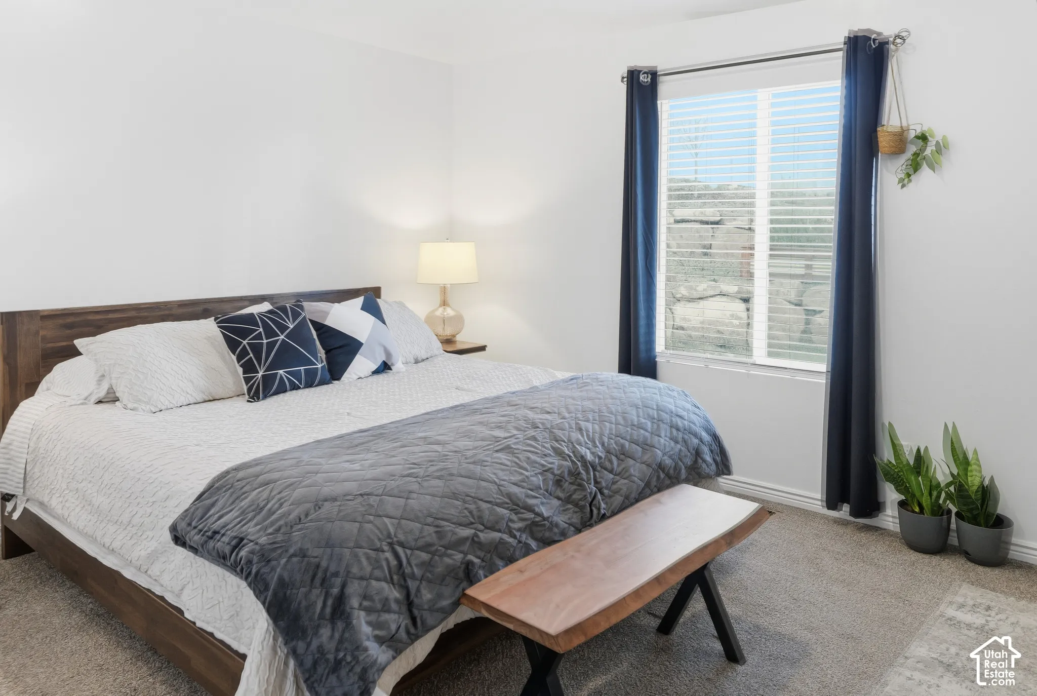 Carpeted room with a large window featuring blinds and dark drapery, accompanied by a built-in shelf and a table lamp