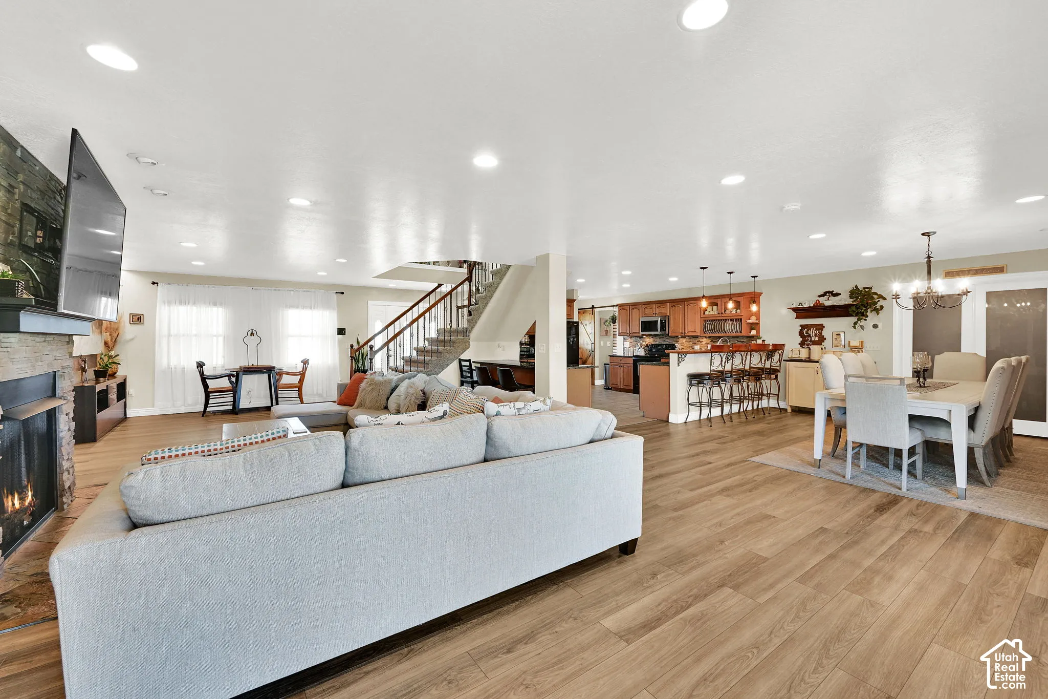 Open-concept living space featuring a light gray upholstered sectional sofa, wood-finish flooring throughout, recessed ceiling lighting, and a staircase with black metal railings