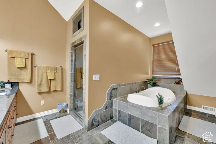 Built-in soaking tub with surrounding tile, glass-enclosed shower with tile surround, dual vanity with stone countertop, and tile flooring