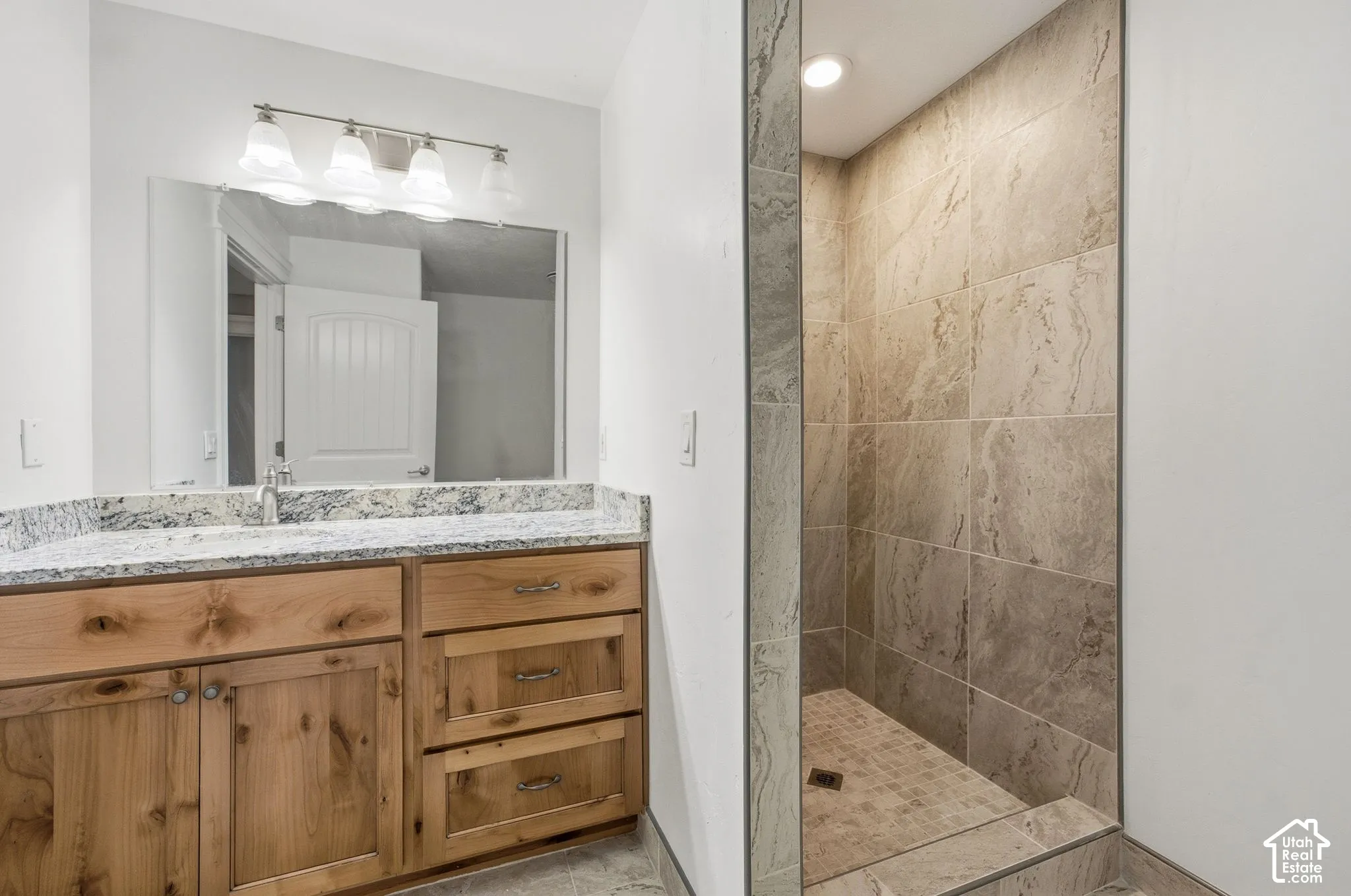 Walk-in shower with tiled walls and floor, wood-finish vanity with a light-toned countertop, and an overhead vanity light fixture