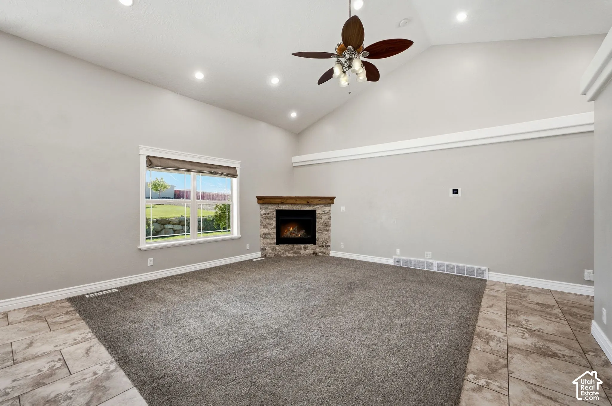 Vaulted ceiling with recessed lighting, ceiling fan with light fixture, stone-faced fireplace with wood mantel, large window with roller shade, and mixed flooring with carpet and tile