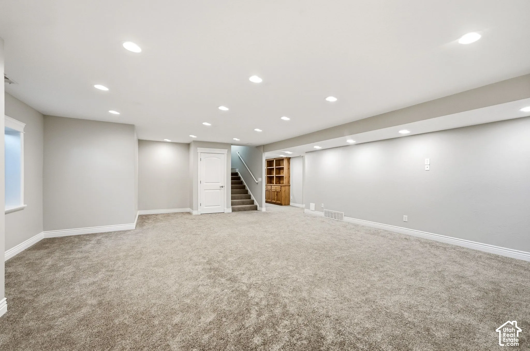 Expansive carpeted room with recessed lighting, integrated wooden shelving, and a stairway with a handrail