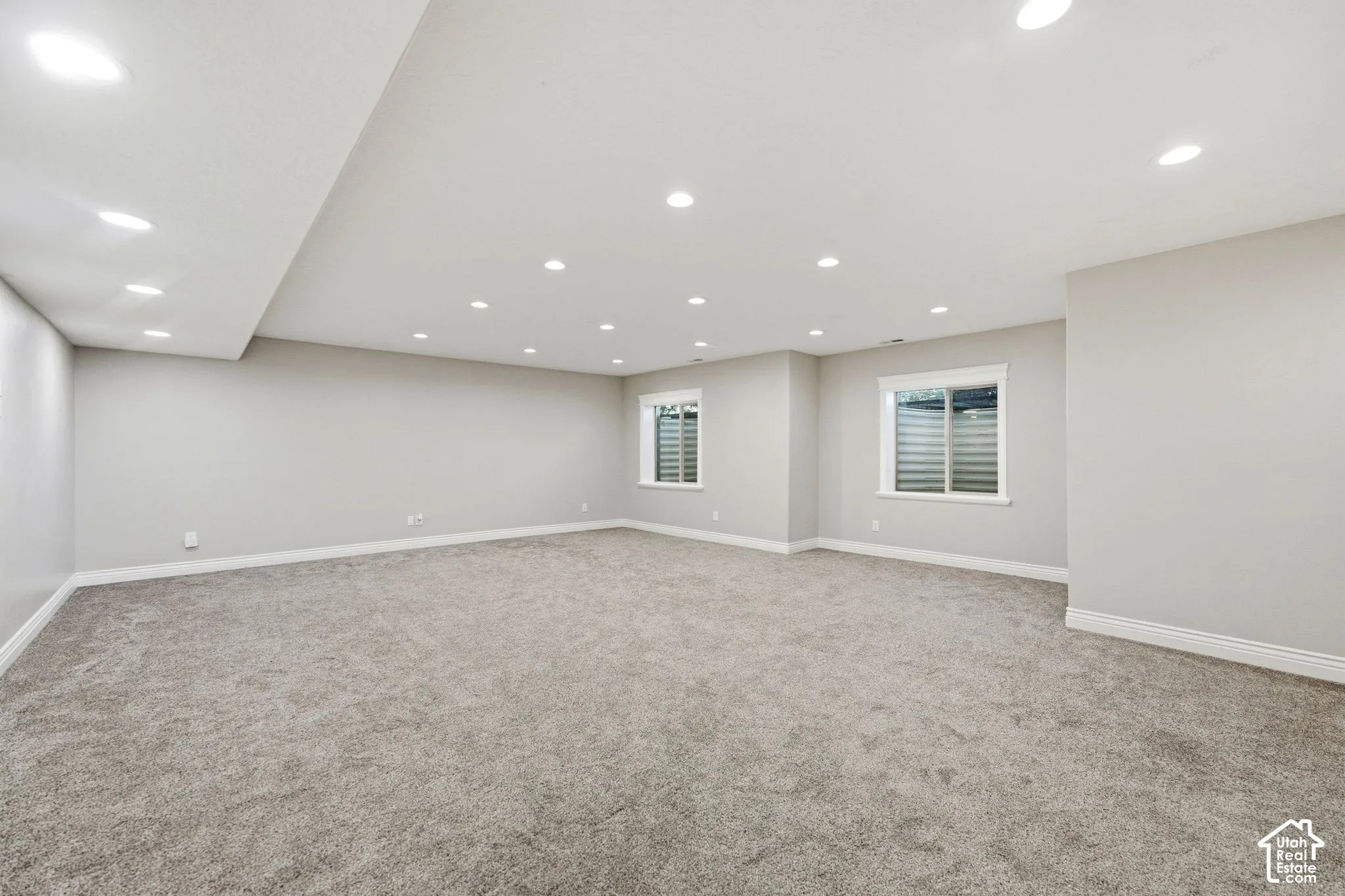 Expansive carpeted area with recessed lighting, light gray walls, and two windows with white trim