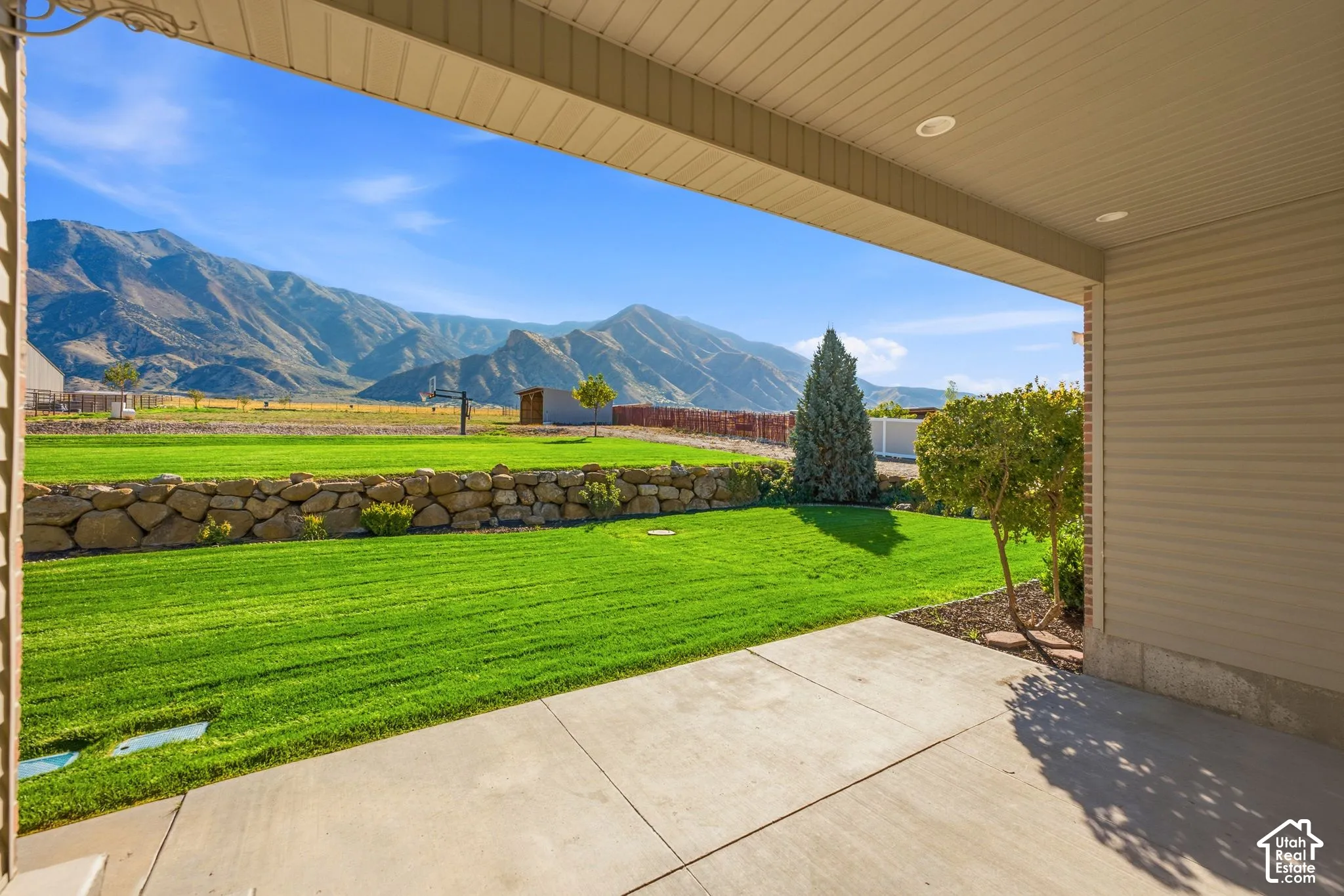 Covered patio with recessed lighting, concrete surface, expansive green lawn, stone retaining wall, and mountain backdrop