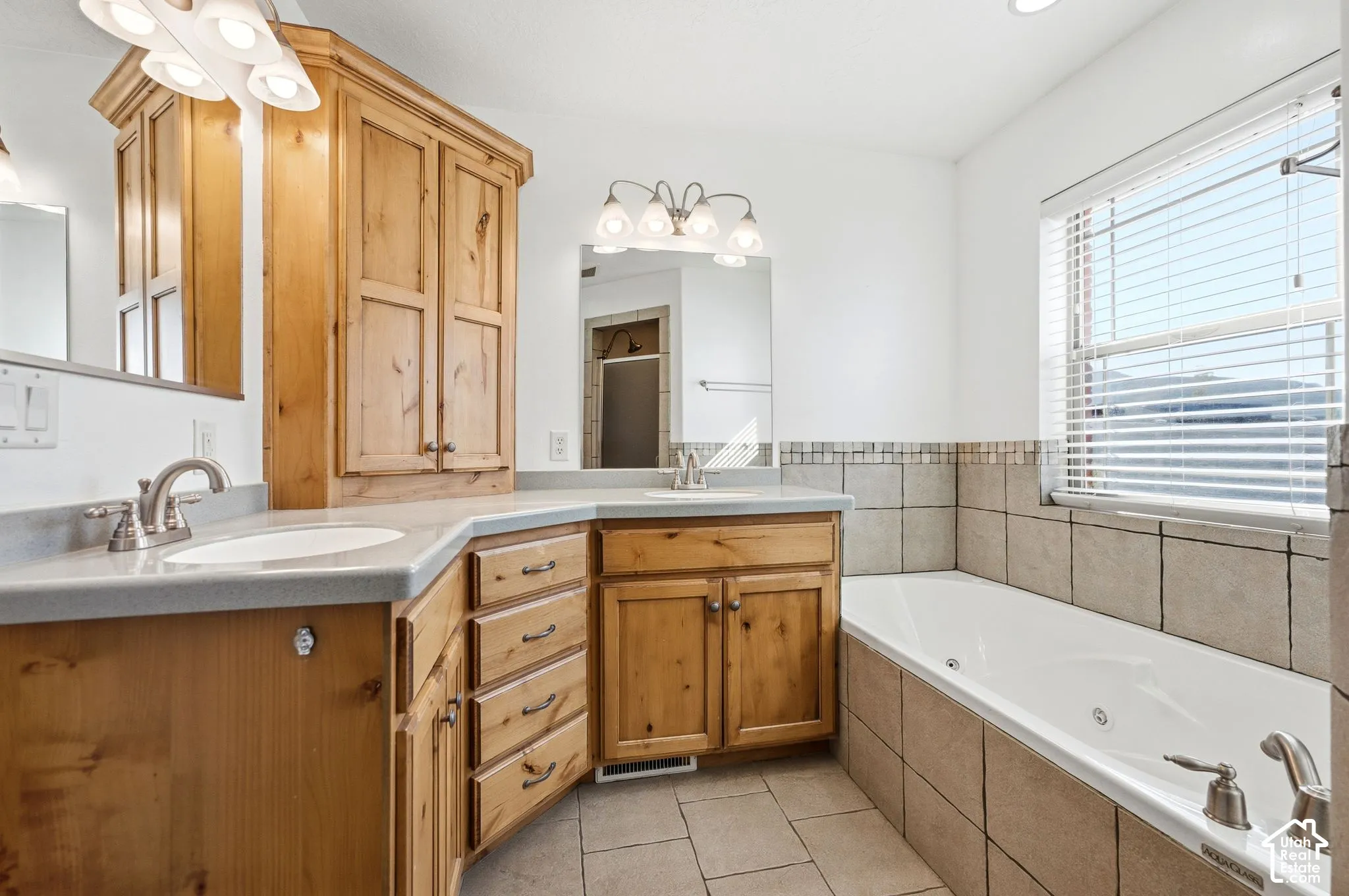 Dual vanity with light wood cabinetry and integrated sinks, built-in storage cabinet, tiled flooring, and a large soaking tub