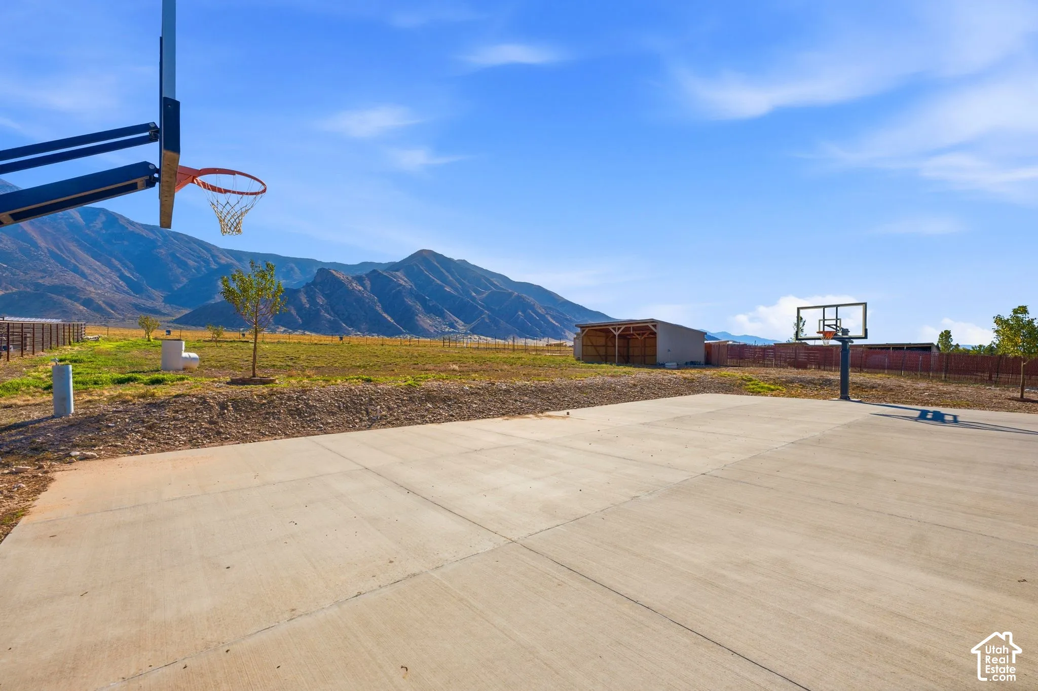 Expansive concrete court with two basketball hoops, a detached shed, a perimeter fence, and a backdrop of mountains