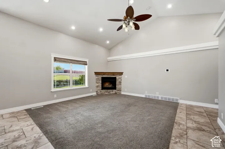 Vaulted ceiling with recessed lighting, ceiling fan with light fixture, stone-faced fireplace with wood mantel, large window with roller shade, and mixed flooring with carpet and tile